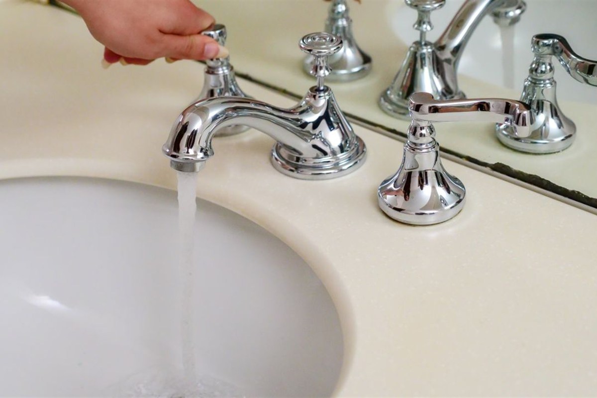 A hand turns on a bathroom faucet with water flowing into the sink.